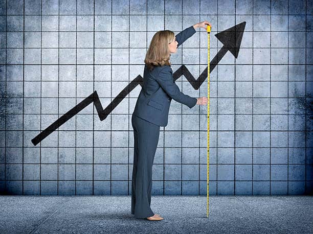 A businesswoman uses a tape measure to measure the growth of an upward pointing graph projected on a concrete wall.