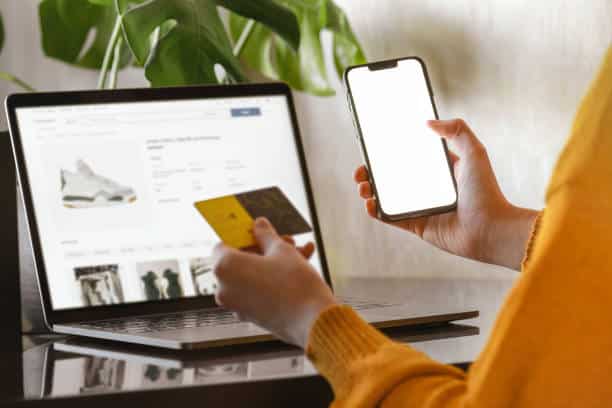Female hands in yellow sweater Setting up her online store  using a laptop, credit card, and mobile phone with white blank screen.